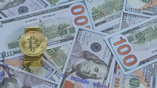 Hand places gold bitcoin coin onto solid gold bar over bed of US hundred-dollar bills