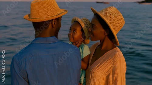 Family by the water at golden hour wearing straw hats with small child in parents arms for summer vacation background and lifestyle banner