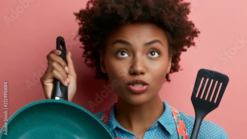 Surprised woman holding a teal frying pan and spatula in floral apron on pink background for kitchen banner and cooking blog design