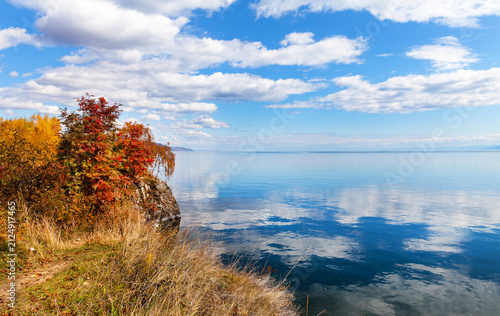 Beautiful autumn landscape of Baikal Lake. View from Cape Shamansky to bay with reflection of clouds in blue calm water. Red rowan bushes on shore at sunny day. Nature background. Travel and outdoors