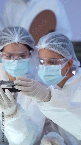 Female engineer team inspecting electronic circuit board component while wearing cleanroom protective suit hairnet face mask and safety goggle inside facility.
