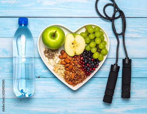 Overhead shot of heart-shaped plate with fruit, nuts, water bottle & jump rope on blue wood