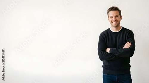 Professional portrait of a smiling mid-adult Caucasian man with arms crossed, wearing a black ribbed sweater and blue jeans, standing against a clean white background with copy space.