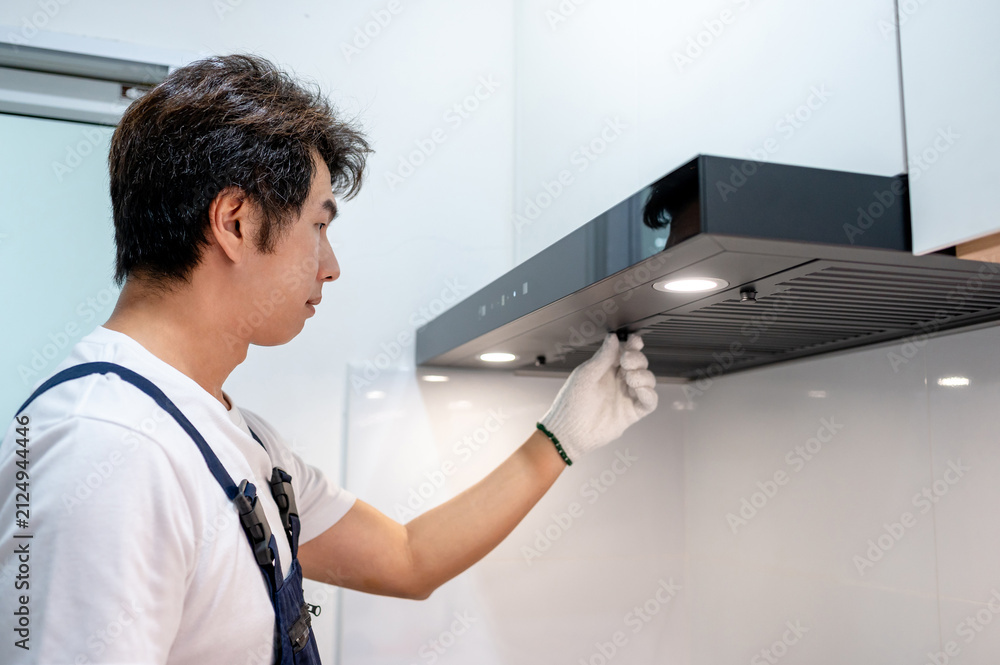 Obraz premium Asian male maintenance worker repairing modern cooker hood in domestic kitchen. Technician man wearing gloves cleaning kitchen range hood filter for clear ventilation. Home appliance service on site