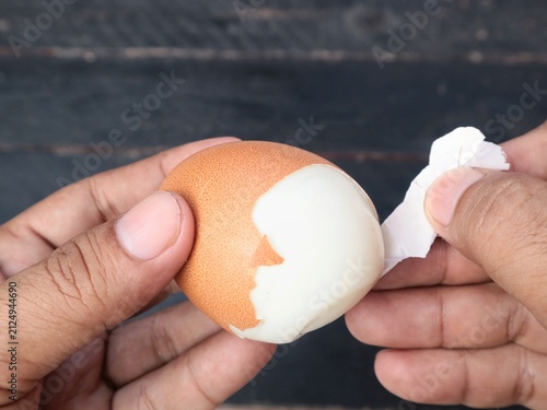 Close up view of human hands peeling the shell of a hard boiled brown chicken egg over a dark wooden background. Concept of healthy breakfast preparation, cooking, protein food, and kitchen routine