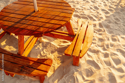 round wooden picnic table with attached benches resting on sunlit sand beside calm beach shoreline in warm