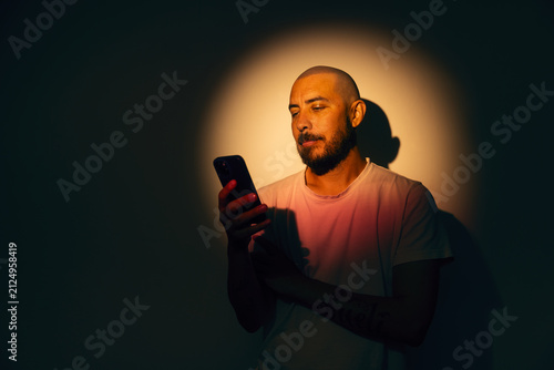 Bald man with a beard looking intently at a smartphone in a dark room illuminated by a warm round spotlight, holding a mobile device with warm lighting and shadow backdrop, expressing focused emotion.