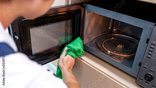 Male hand holding green microfiber cloth wiping inside the black microwave oven in wooden cabinet. Cleaning home appliance in the kitchen