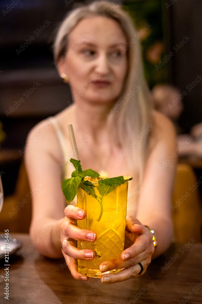 Fototapeta premium Woman extends a glass of non alcoholic mojito with lime, mint and ice toward the camera in a restaurant. Invitation to refreshment concept supporting hospitality, celebration and customer experience.