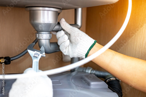 Plumber hand with glove fixing pipe connection with adjustable wrench and checking grease trap box under kitchen sink. Handyman repairing plumbing system in wooden cabinet.