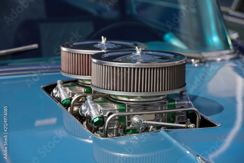 Close-up of a classic vintage car engine compartment featuring a dual-quad carburetor setup with custom chrome air cleaners protruding through a cut-out in the blue hood