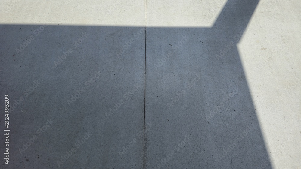 Fototapeta premium Smooth concrete floor with sharp sunlight and line shadows. High-angle view of a grey cement ground with dramatic morning sun rays and architectural shadows.