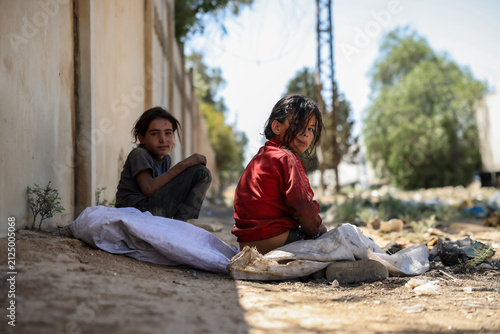 Thoughtful poor children sitting near a wall. Emotional portrait of a young refugee girl with sad eyes. Two Syrian girls in torn clothes sit by the roadside after rummaging through garbage for plastic