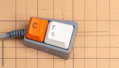 Small two‑key mechanical keyboard placed on grid‑patterned surface, minimalist keypad design with one orange key labeled “C” and one white key labeled “T Q,” cable attached suggesting connection to co