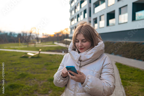 A woman is smiling and looking at the camera in a grassy outdoor area with a modern building in the background. The scene conveys a happy and relaxed mood, with natural green and white hues.