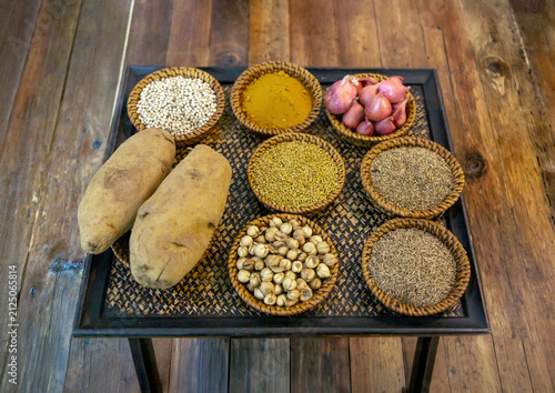 assortment of various aromatic spices, seeds, fresh shallots, and potatoes arranged in wicker bowls on a rustic wooden table for cooking preparation.