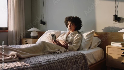 Camera pulling back revealing woman wearing robe, holding smartphone on bed and getting comfortable