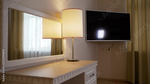 Warm lit hotel room corner featuring wooden desk, white framed mirror, table lamp with cream shade. Soft ambient lighting, neutral tones, modern decor