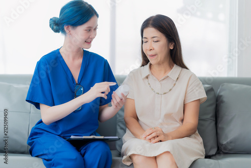Young Asian female doctor or nurse with blue hair explaining dosage instructions for medication bottle to a senior female patient in a hospital or clinic.