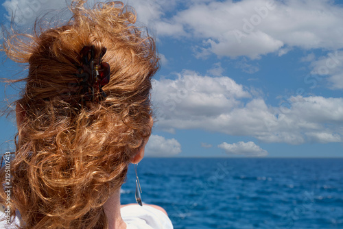 Obraz na plátne Back view of woman with curly reddish hair looking out at blue sea and cloudy horizon in Gran Canaria