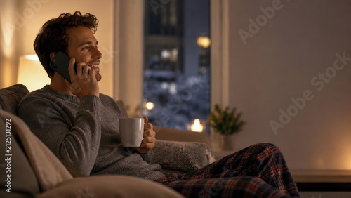 Relaxed young man sitting on comfortable sofa at night, smiling while talking on his mobile phone and holding warm mug of coffee in cozy dimly lit home interior setting.