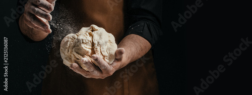 Male baker wearing brown apron holds kneaded dough with flour dusting in a dark kitchen setting, showcasing hands and texture of the dough