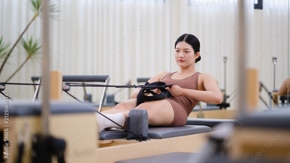 Fototapeta premium Young woman exercising on a Pilates reformer machine with resistance straps in a bright modern fitness studio.