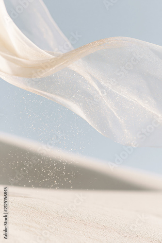 Translucent ivory fabric billows above pale sand, scattering shimmering particles against a softly blurred blue sky. Selective soft focus.