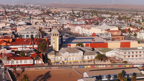 Aerial view of Swakopmund, Namibia, with German era architecture, yellow and white tower, murals on the seawall, palm lined beach, and Namib Desert dunes in warm light.