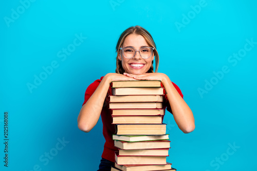 Fotografía Happy female student with glasses leaning on a tall stack of book against a blue