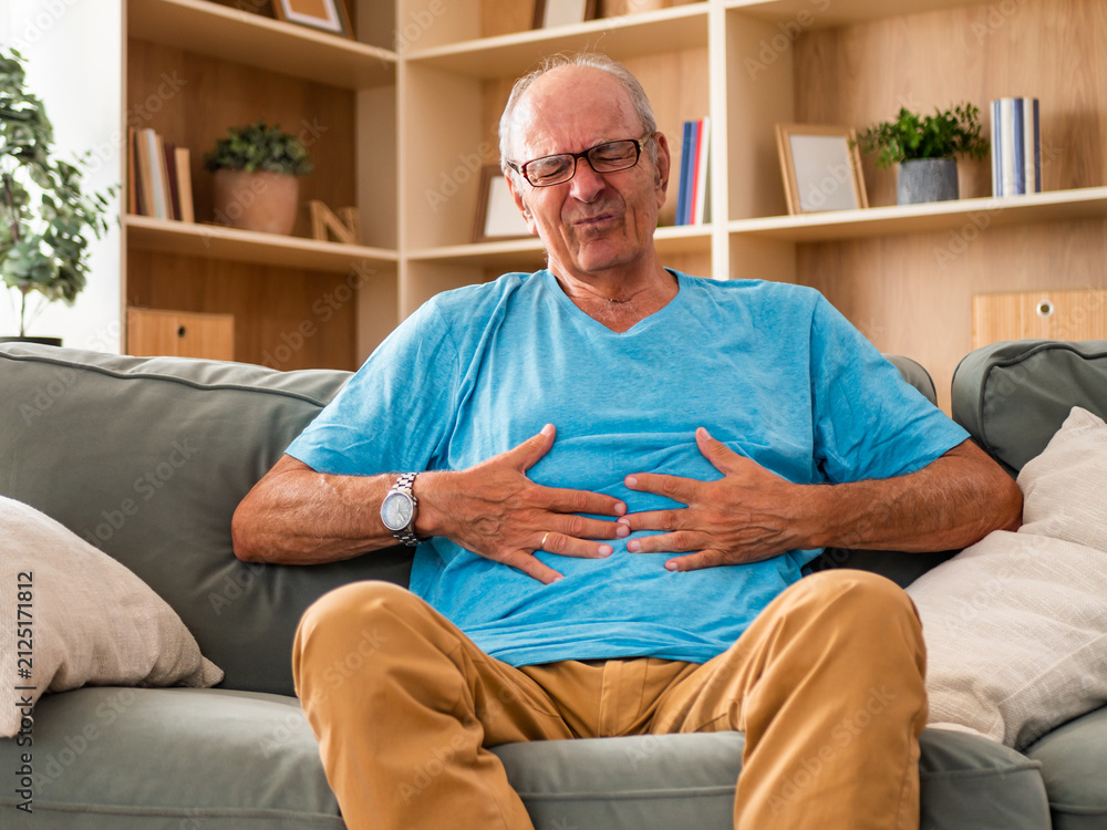 Fototapeta premium Senior man sitting on sofa in his living room and holding his abdomen with a painful expression. He is illustrating stomachache, indigestion, digestive problems and discomfort at home.