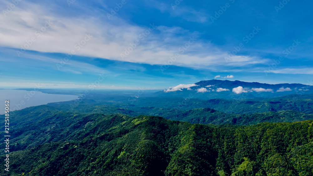 Fototapeta premium Mountain ranges stretch to the distant coast beneath a wide blue sky. Layers of green hills and drifting clouds create a dramatic tropical aerial landscape.