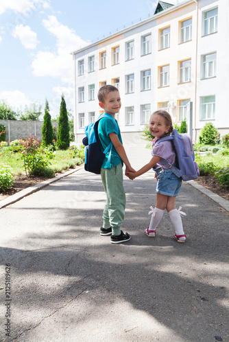 Two smiling elementary school children with backpacks holding hands while walking toward the school building on a sunny morning
