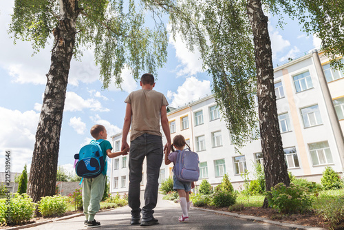 Caring father walks hand in hand with his young son and daughter toward the school entrance on a sunny back to school morning