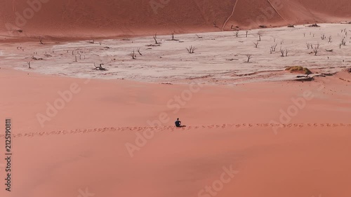 Aerial view of Deadvlei in Sossusvlei, Namib Desert, Namibia, with rust red dunes, dead camel thorn trees, a lone person on a ridge, and tiny figures near the pan.