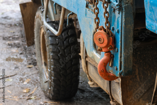 Old industrial tow hook hanging from weathered blue truck with mud covered tire and chain, gritty worksite atmosphere conveying rugged utility and worn texture