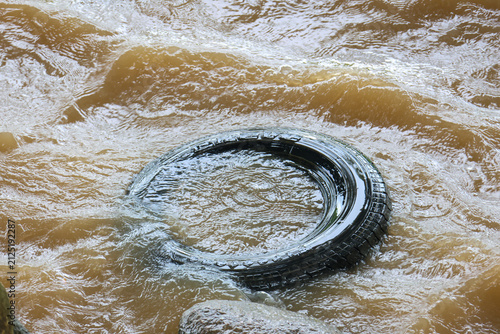 Old rubber tire partially submerged in muddy flowing river water, pollution scene with brown current and textured ripples conveying environmental impact and neglect