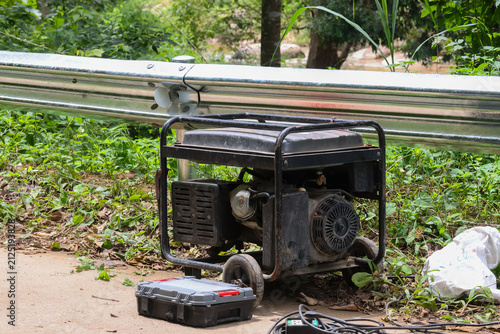 Portable generator on grassy roadside with repair toolbox and cables, rugged metal frame and wheels, outdoor construction equipment providing emergency power for worksite or camping