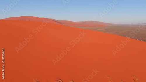 Aerial view of a towering orange red dune in Sossusvlei, Namibia. Small groups walk the knife edge crest as a sweeping low shadow moves in warm morning or late day light.