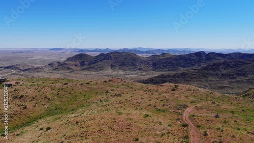 Aerial view over Namibia Damaraland shows rocky hills, broad plains, and a meandering dirt track at midday. Layered ranges recede, sparse shrubs and ochre grasses show depth.