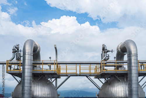 Two large insulated pressure vessels connected by arched metal pipes and a yellow walkway at an LNG or gas processing plant with mountains and blue sky in the background showcasing industrial tank inf