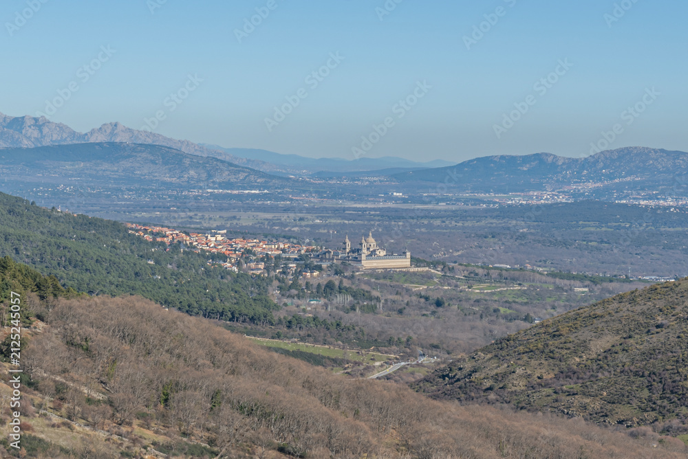Fototapeta premium Panoramic view of El Escorial monastery and town nestled in a valley surrounded by forested hills and distant mountains.