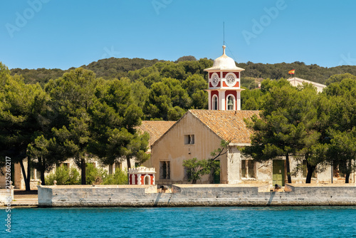 Red and white clock tower and old building surrounded by Mediterranean sea and lush greenery in the port of Mahon, Spain