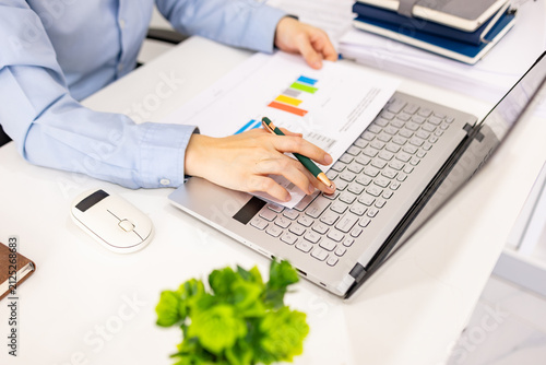 Woman analyzes charts on a laptop. She studies data with focused intent. Desk holds plants, notes, and a phone. Professional workspace with soft lighting
