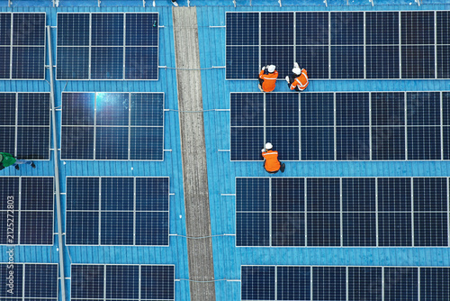 Aerial view of technicians installing solar panels on blue roof. Engineers maintaining solar energy system on a building rooftop. Top down view of workers in orange uniform fixing solar panels. 