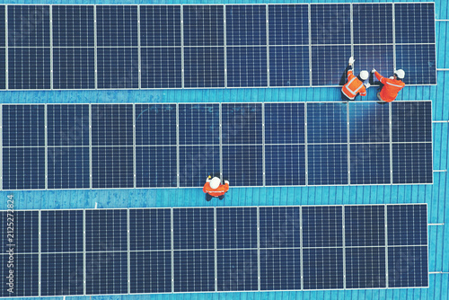 Aerial view of technicians installing solar panels on blue roof. Engineers maintaining solar energy system on a building rooftop. Top down view of workers in orange uniform fixing solar panels. 