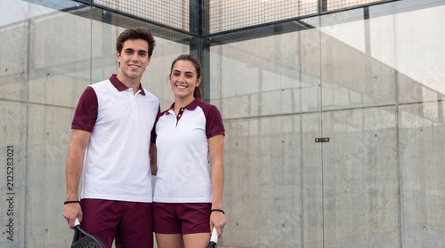 Two players in matching uniforms holding padel rackets on glass court