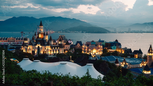 Illuminated castle-inspired of Vinpearl in VinWonders theme park overlook a scenic bay at dusk, creating a magical waterfront cityscape with mountains, dramatic clouds, and evening lights.