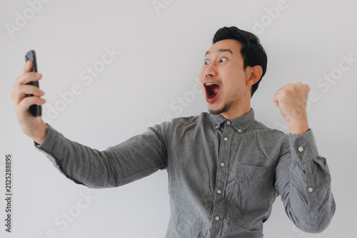 Obraz na plátne Excited Asian man celebrating success while looking at his smartphone with a triumphant fist