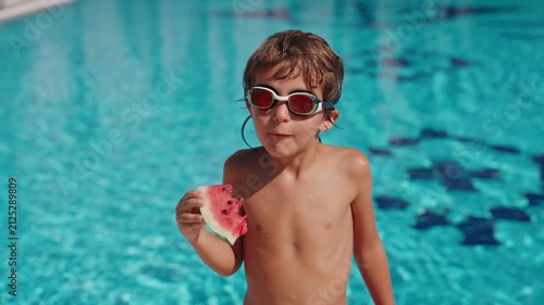 A smiling boy wearing swimming goggles enjoys a fresh slice of watermelon by an outdoor swimming pool on a bright summer day. The child stands poolside after swimming, eating juicy seasonal fruit